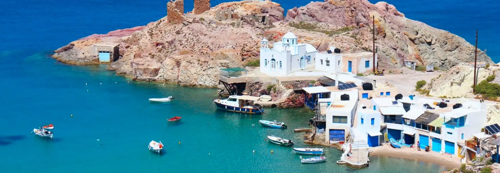 Aerial view of Firopotamos bay with fishing boats on turquoise water, white chapel, and blue-door boat houses along rocky cliffs