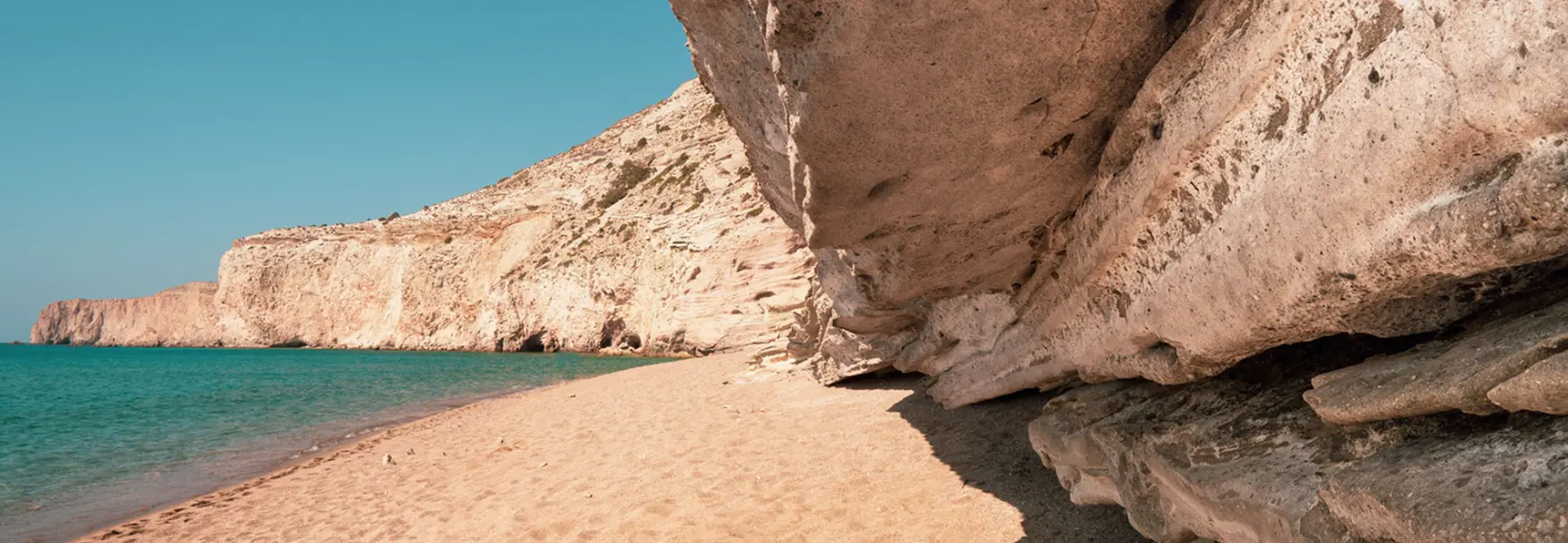 Sandy beach beneath eroded volcanic rock overhangs with turquoise water at Gerakas in Milos