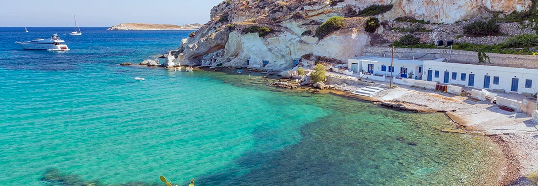 Turquoise waters of Rema cove on Kimolos with traditional white fishing houses and a Navy Blue yacht anchored offshore