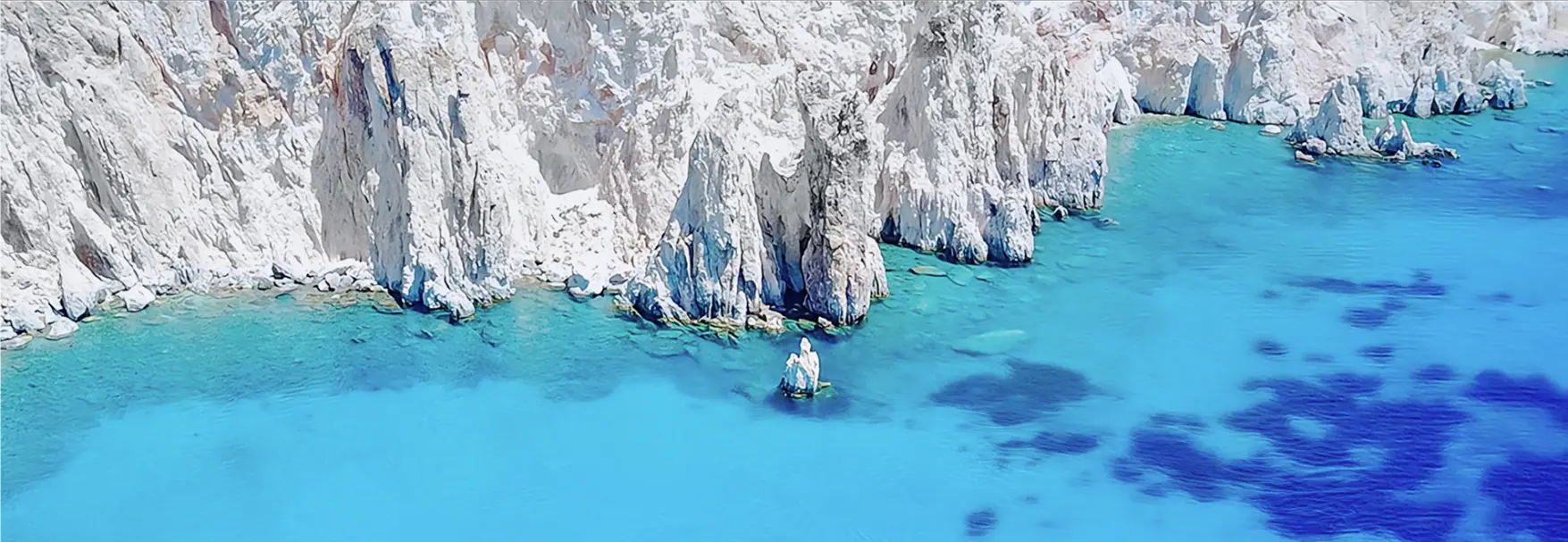 Aerial view of jagged white rock formations rising from crystal-clear turquoise waters along the Poliegos coastline