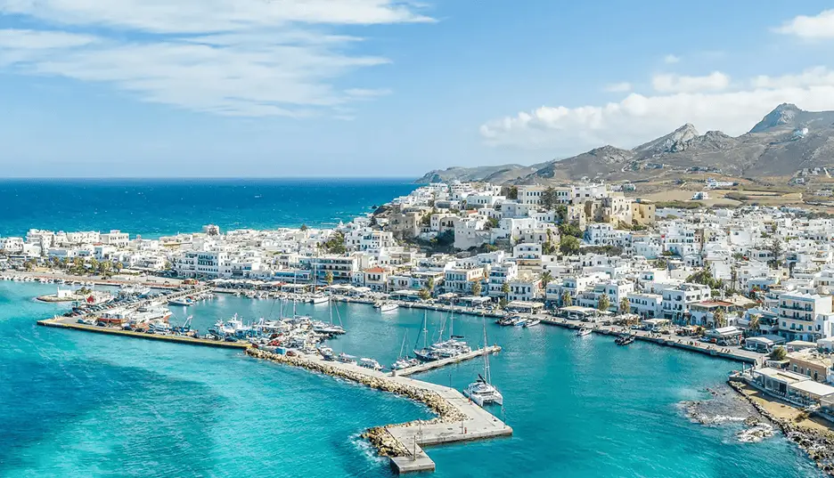 Aerial view of a Cyclades island town with a yacht-filled marina, whitewashed buildings, and mountain backdrop