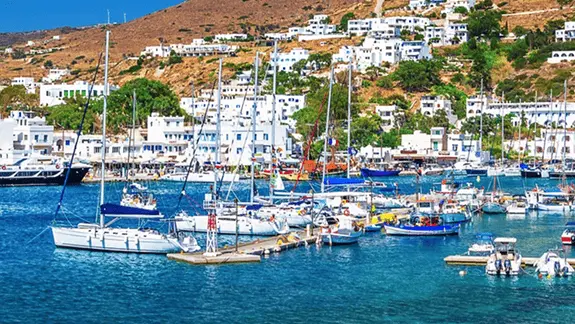 Sailboats moored in a Cyclades island marina with whitewashed village and green hillside above