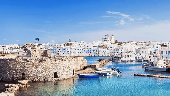 Stone medieval castle with Greek flag above a Cyclades harbor with small boats and white cubic buildings