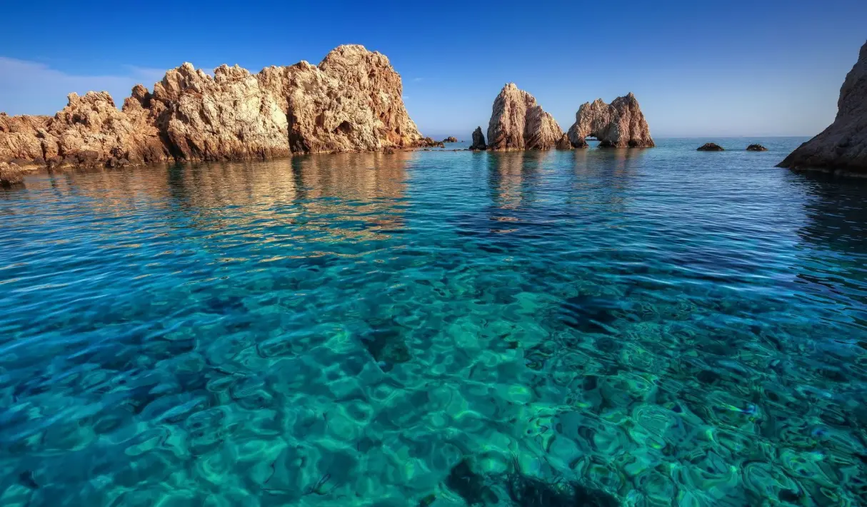 Crystal-clear turquoise water over the rocky seabed near Antiparos with jagged sea stacks rising from the calm surface under a blue sky