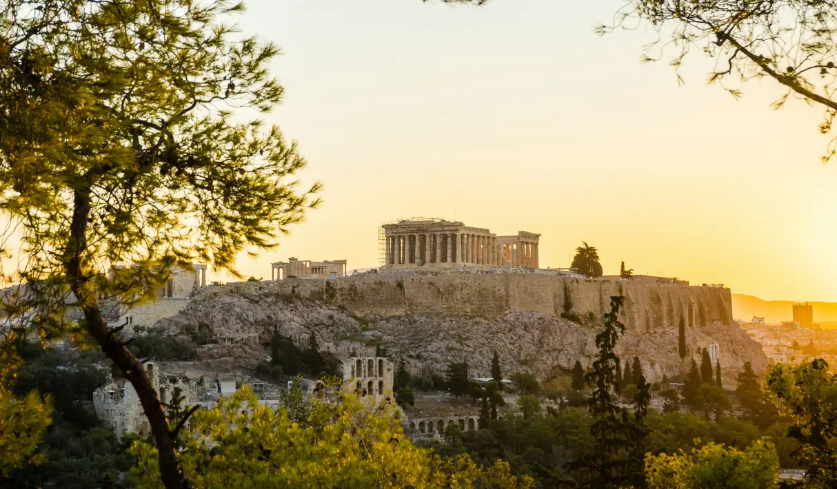 The Acropolis of Athens with the Parthenon atop a rocky hill at golden sunset, framed by trees with the Odeon of Herodes visible below