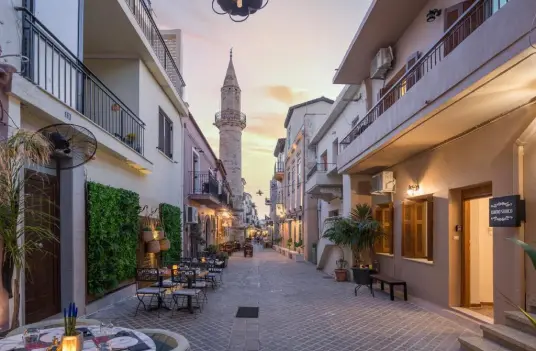 A narrow street in the old town of Chania at dusk, with lit tavernas and a minaret above the roofs