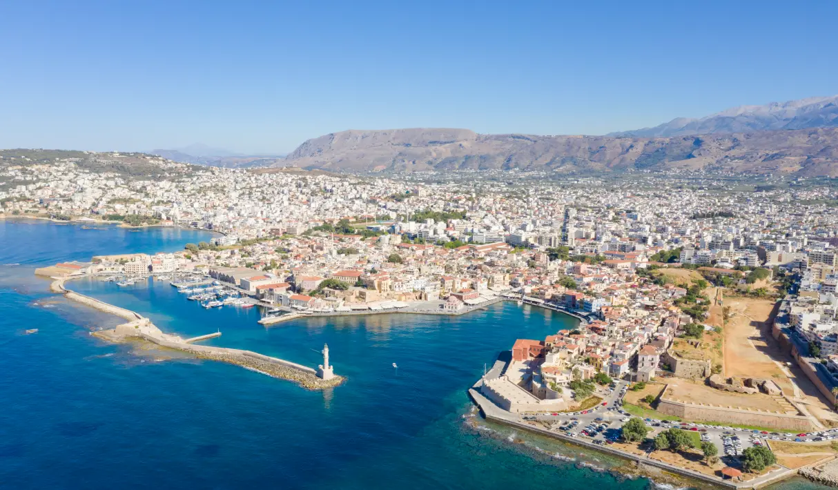 Aerial view of Chania, Crete, with a Venetian lighthouse at the harbor entrance, old fortress walls, and the city extending to mountain ranges