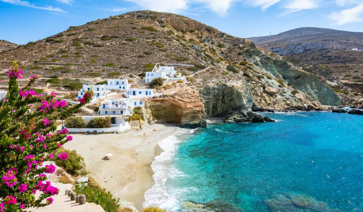 Secluded sandy beach in Folegandros with turquoise water, whitewashed cliffside buildings, a rocky headland, and pink bougainvillea