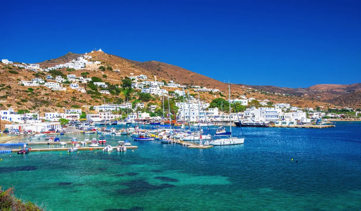 Busy Ios marina packed with sailboats and yachts moored on blue water with whitewashed buildings climbing the hillside behind