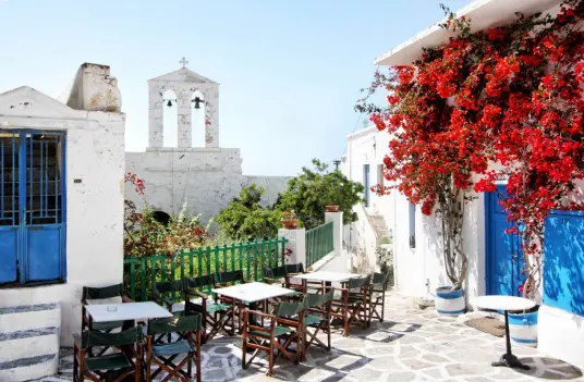 A whitewashed cafe terrace on Kimolos with a bell tower, blue shutters, and red bougainvillea
