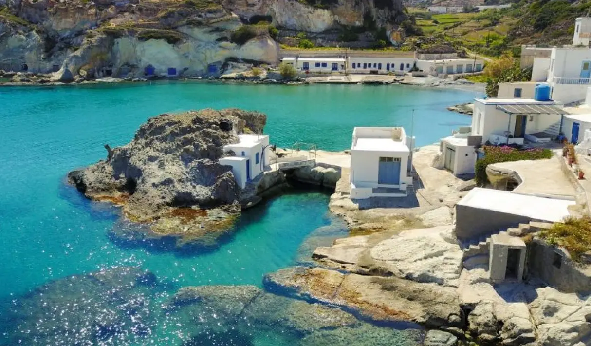White fishermen's houses built directly onto the rocky shoreline of Kimolos with a large sea rock and vivid turquoise water