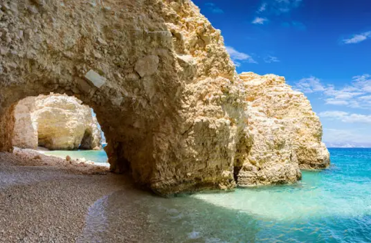 A wide sea cave arch in pale rock opening onto a small beach and turquoise water on Koufonisia