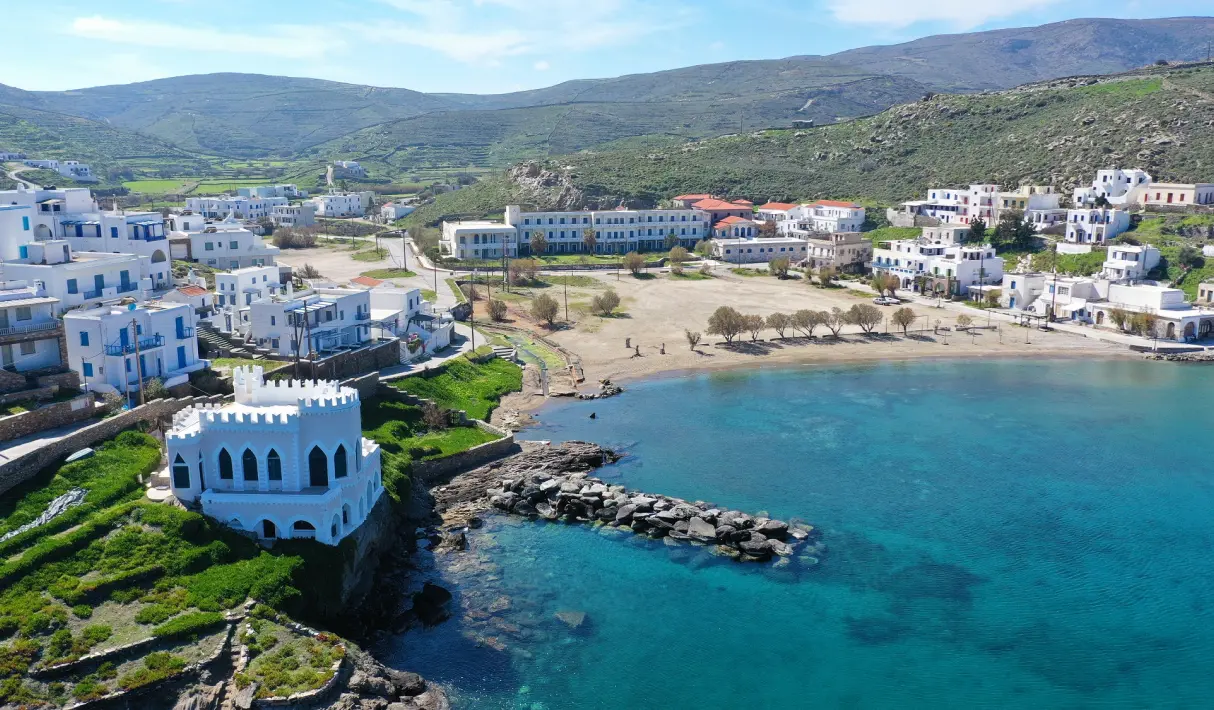 Aerial view of Kythnos coastal village with a white crenellated waterfront building, a sandy beach, turquoise water, and green rolling hills