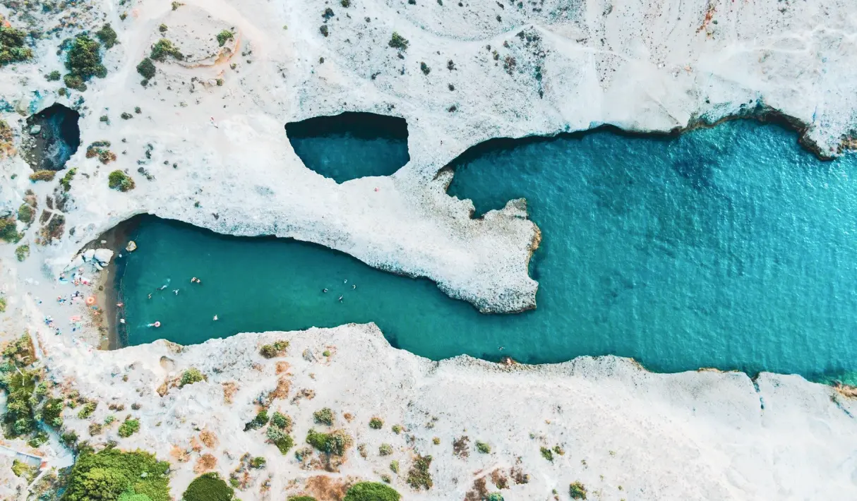 Aerial view of white volcanic cliffs and a turquoise cove on Milos in the western Cyclades, by Navy Blue