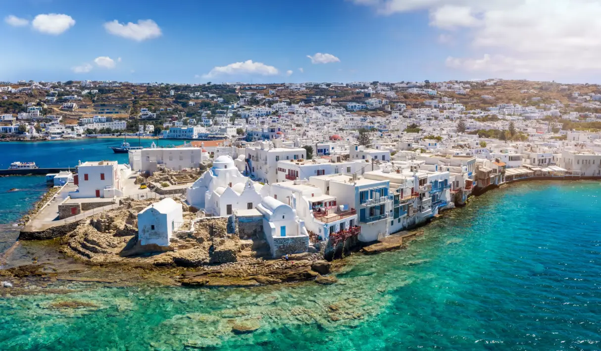 Aerial view of Mykonos Chora with hilltop windmills, white waterfront buildings, a sandy beach, and a sailing vessel in the harbor