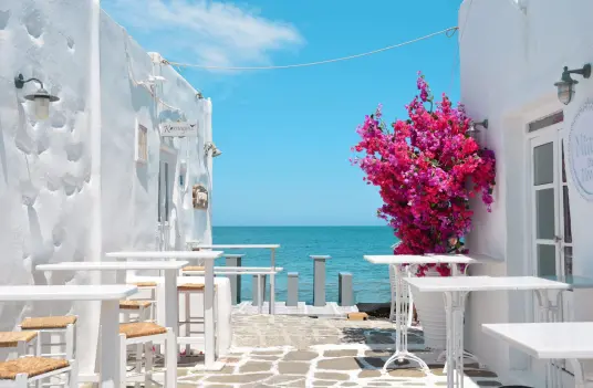 Whitewashed Cycladic alley on Paros with cafe tables, bright bougainvillea, and an Aegean Sea view at the end
