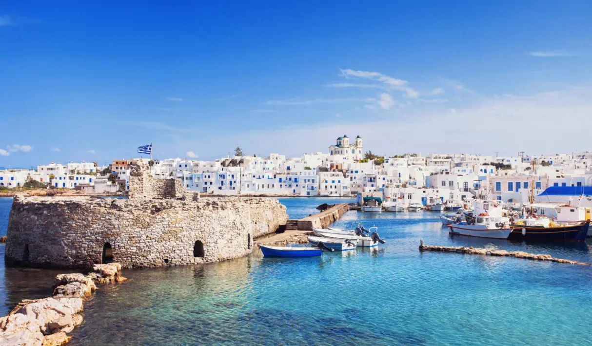 Ancient stone harbor fortress in Paros with moored fishing boats, whitewashed waterfront buildings, and a Greek flag under a deep blue sky