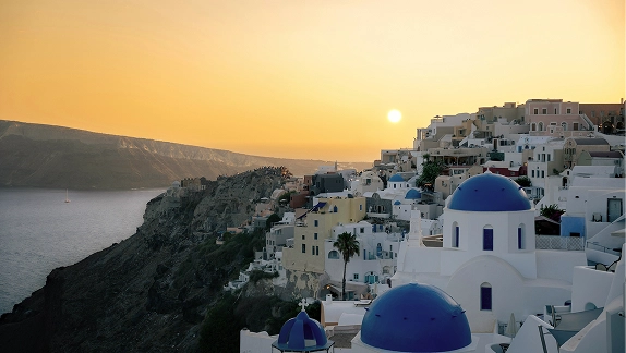 Golden sunset over Santorini's Oia with terraced white buildings and a blue-domed church on the clifftop