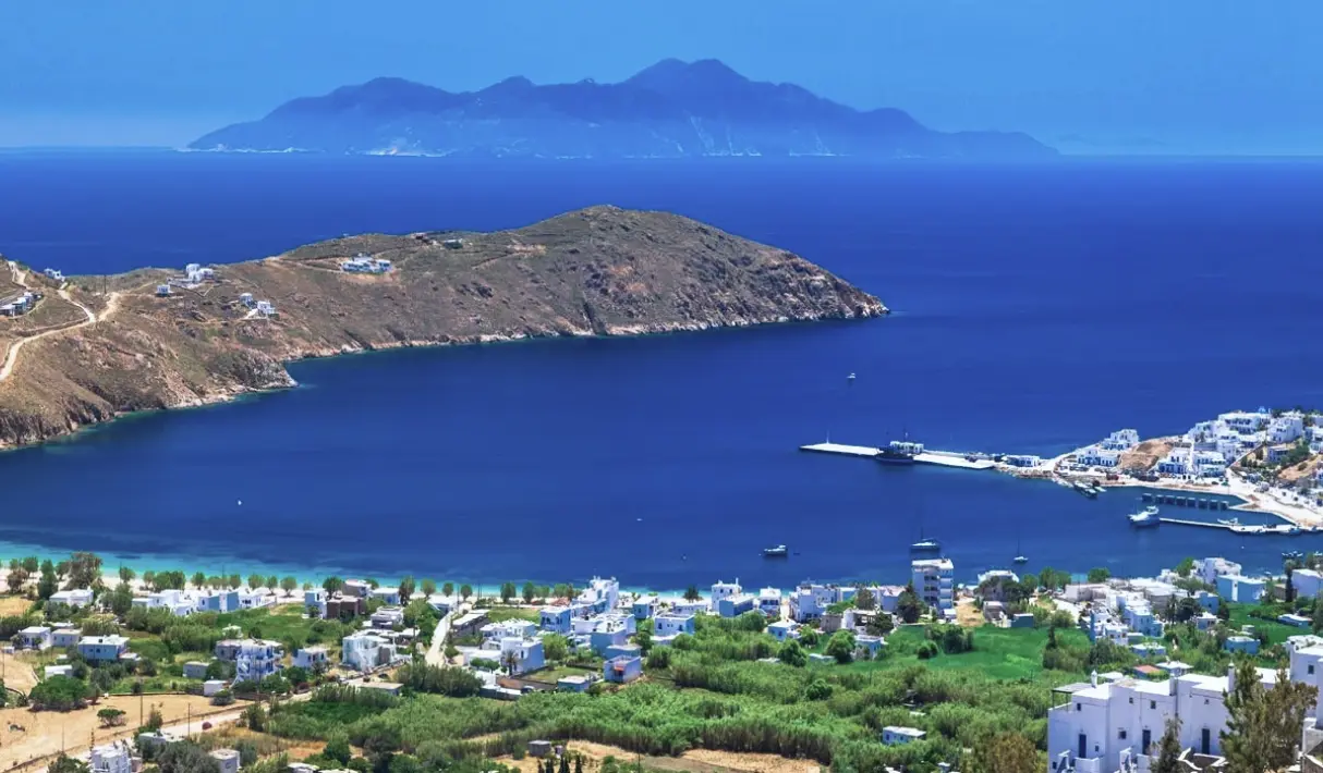 Aerial view of Serifos bay with a green coastal town below, a rocky headland extending into deep blue sea, and a distant island silhouette