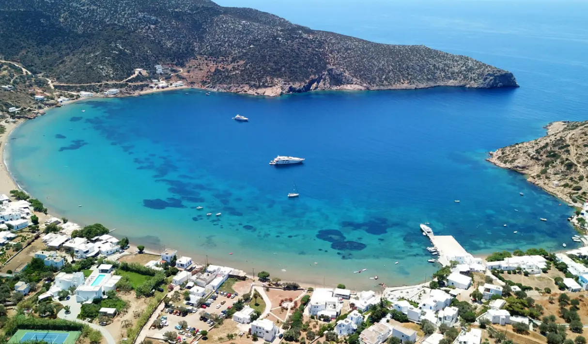 Aerial view of a sheltered circular bay in Sifnos with two yachts moored on turquoise water, a sandy beach, and a rocky scrub hillside