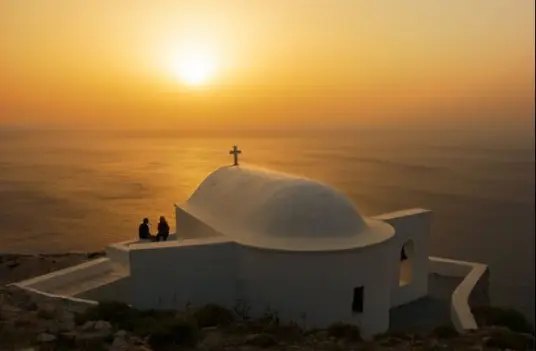 A domed whitewashed church on a clifftop above the Aegean on Sikinos, with two people at sunset