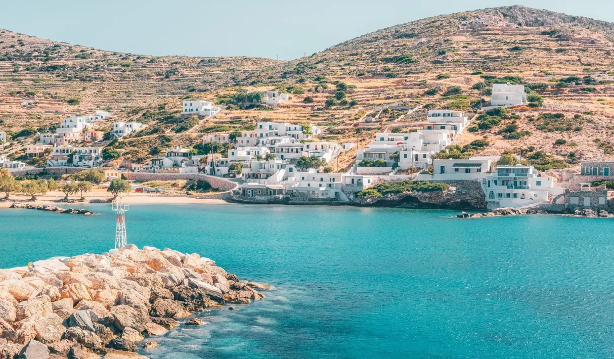 Sikinos small harbor with a stone breakwater, a white lighthouse beacon, whitewashed hillside buildings, and calm turquoise water