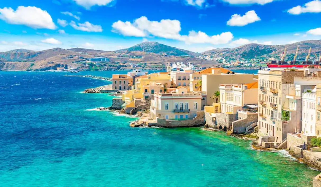 Neoclassical ochre and yellow buildings of Syros built directly along a rocky turquoise waterfront with stone sea walls and palm trees