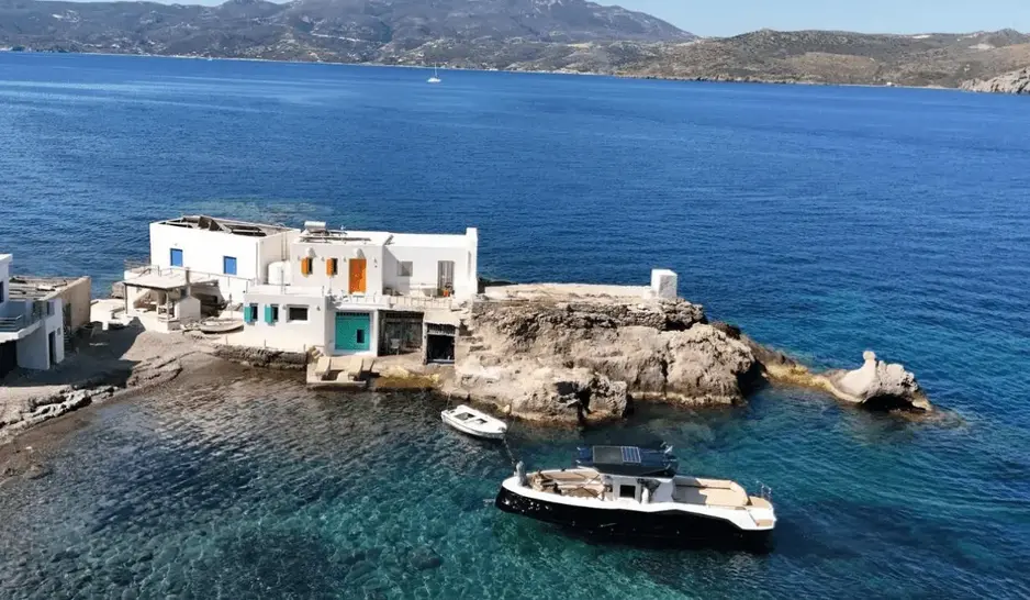 Aerial view of a Navy Blue yacht anchored beside traditional Milos stone houses on the crystal-clear coastline
